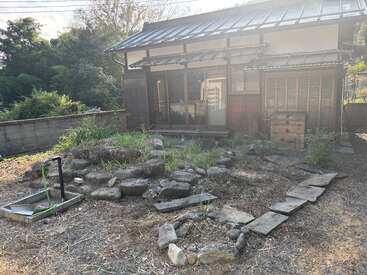 Uma casa tradicional japonesa com telhado de telha fica em meio à vegetação. O jardim tem pedras, um caminho de pedra, um pouco de grama e uma área de pia externa.