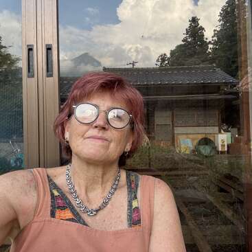 A woman with short red hair and glasses takes a selfie outside. Reflections show clouds and scenery behind her. She wears a colorful top and a chain necklace.