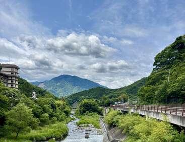 Um rio flui entre árvores e colinas verdejantes, com uma estrada e um prédio de cada lado, sob um céu azul parcialmente nublado e montanhas.