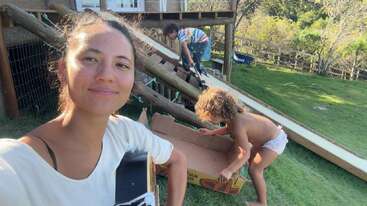 Una mujer sonríe a la cámara al aire libre, con una guitarra en la mano. Dos niños juegan cerca: uno dentro de una caja y otro subiéndose a un tobogán. Es una escena alegre y llena de alegría.