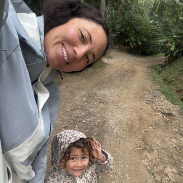 Una mujer y un niño sonríen en un camino de tierra rodeados de una exuberante vegetación. El niño lleva una chaqueta con capucha estampada; ambos parecen felices al aire libre.