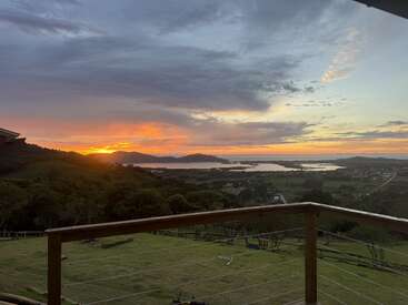 Una impresionante puesta de sol ilumina el horizonte, bañando con cálidos colores las onduladas colinas, un tranquilo lago, los árboles y las casas, todo ello contemplado desde un balcón de madera con barandilla de alambre.