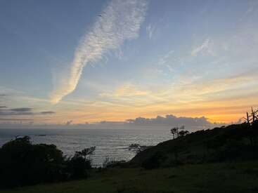 Una tranquila escena costera al atardecer, con un horizonte de un naranja resplandeciente, nubes espectaculares, un mar en calma, árboles recortados contra el cielo, colinas cubiertas de hierba y un pájaro volando en lo alto.