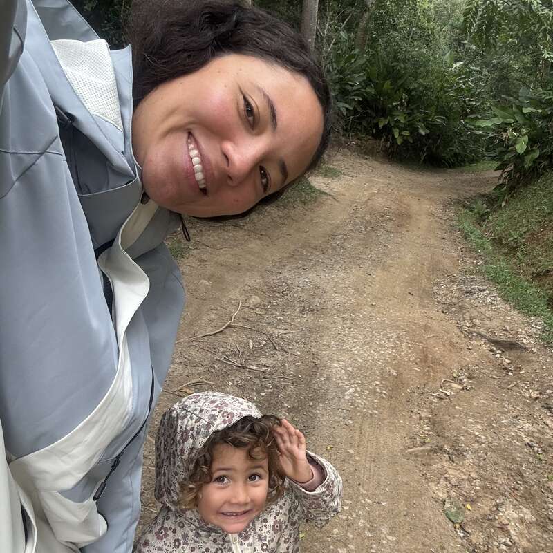 Una mujer y un niño sonríen en un camino de tierra rodeados de una exuberante vegetación. El niño lleva una chaqueta con capucha estampada; ambos parecen felices al aire libre.