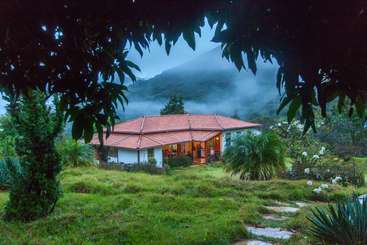 A imagem retrata uma cena serena de uma casa com um grande quintal, com uma árvore e uma cerca, tendo como pano de fundo um céu claro e um corpo d'água.