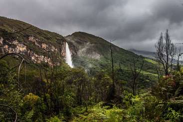 A imagem retrata uma cachoeira majestosa, com uma grande massa de água caindo em cascata em um penhasco rochoso, cercada por uma vegetação exuberante e um céu azul claro. A cena exala beleza natural e tranquilidade.
