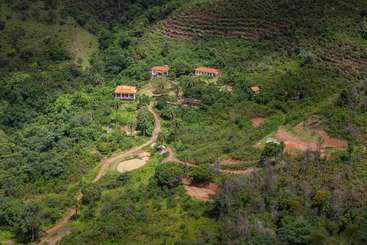 A imagem retrata uma paisagem serena com um conjunto de edifícios, provavelmente casas, em meio a uma vegetação exuberante, com uma estrada sinuosa e um corpo d'água, evocando uma sensação de tranquilidade e beleza natural.