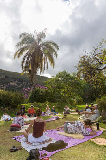 A imagem retrata uma reunião serena de pessoas ao ar livre em uma colina gramada, cercada por árvores exuberantes e uma palmeira majestosa, com um céu nublado acima.