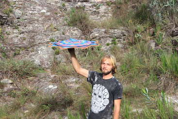 Uma pessoa está em uma praia, segurando uma pipa, com um corpo de água e dunas de areia ao fundo, criando uma atmosfera serena e pacífica. O céu está limpo e azul.