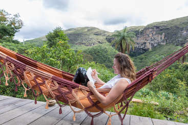 Uma mulher sentada em uma rede, cercada por um ambiente externo sereno, com um corpo de água e vegetação exuberante, evocando uma sensação de relaxamento e tranquilidade.