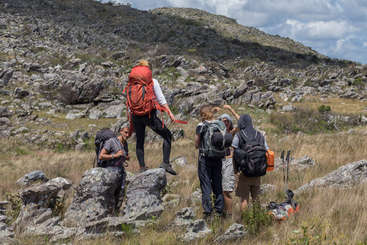 Um grupo de pessoas está caminhando nas montanhas, algumas sentadas em rochas e outras em pé, cercadas por vegetação e um céu claro. A cena é pacífica e serena.