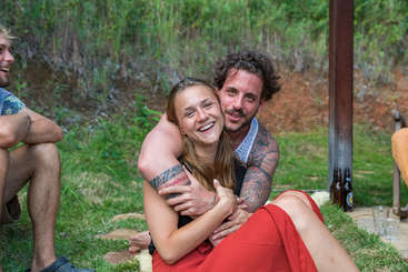 A imagem mostra um grupo de pessoas reunidas, com um homem e uma mulher se abraçando em primeiro plano, cercados por uma multidão de pessoas ao fundo. A cena parece ser uma reunião ou comemoração social.