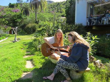 A imagem mostra duas mulheres tocando violão em um dia ensolarado, em um quintal gramado e exuberante, com árvores e uma casa ao fundo, cercadas por uma atmosfera serena.