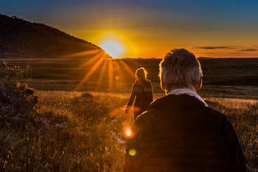Dois indivíduos estão se afastando da câmera, com o homem à esquerda e a mulher à direita, ambos vestindo shorts e camisetas. Eles estão caminhando em uma trilha.