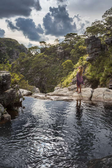 Uma jovem com longos cabelos loiros está em uma pedra à beira de uma piscina em uma área montanhosa verde e exuberante, sob um céu nublado.