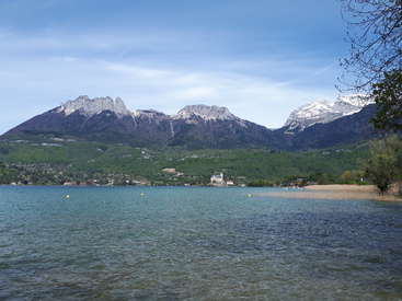 The image depicts a serene lake scene with a majestic mountain range in the background, featuring a small town and a sandy beach along the water\'s edge.
