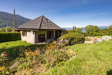 A small cottage with a pyramid-shaped roof sits amidst a lush green garden, surrounded by colorful wildflowers, bushes, and distant mountains under a clear blue sky.