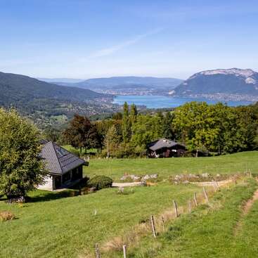 A scenic countryside view features green fields, quaint houses, lush trees, a winding path, a serene blue lake, and surrounding mountains under a clear blue sky.