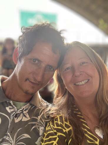 Ein Mann und eine Frau, beide lächelnd, lehnen sich für ein Selfie eng aneinander. Sie tragen gemusterte Hemden und wirken glücklich, während das Sonnenlicht ihre Gesichter sanft beleuchtet.