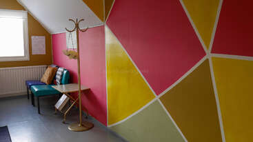 Una sala colorida con motivos geométricos en las paredes en tonos amarillo, rosa y dorado. Las sillas azules y verde azulado, el perchero, la ventana y la mesa crean una acogedora zona de espera.