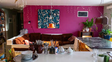 Una sala de estar acogedora y ecléctica con un sofá modular marrón, una pared decorativa en color magenta, arte abstracto, plantas, libros, una mesa de comedor y utensilios de cocina en la encimera.