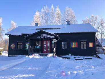 Una casa de madera negra con puertas rojas se encuentra rodeada de nieve, con decoraciones coloridas en sus ventanas y árboles con ramas heladas bajo un cielo azul claro.