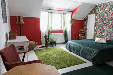 Esta acogedora habitación cuenta con una colcha verde, papel pintado floral, una alfombra verde, paredes rojas, plantas, muebles de madera, un escritorio blanco y una ventana con cortinas verdes.