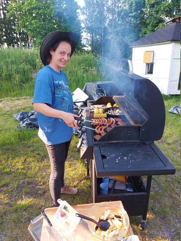 Una persona con un gran sombrero negro y una camisa azul está asando salchichas y maíz al aire libre. Cerca se ven árboles, un cobertizo y una mesa para preparar la comida.