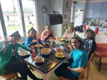 Seis personas están sentadas alrededor de una mesa, sonriendo y con sombreros de hojas. Están disfrutando de una comida juntos en una cocina acogedora y luminosa, levantando las copas con alegría.