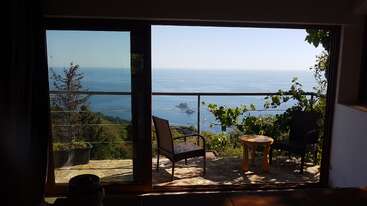 The image depicts a serene outdoor scene, featuring a house with a large window and a patio area, surrounded by a lush green lawn and a tranquil body of water.