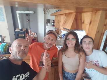 The image shows four people posing for a selfie in a kitchen with wooden cabinets and countertops, giving thumbs up and peace signs.