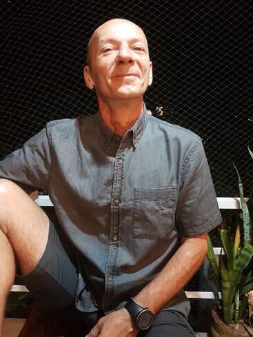 The image depicts a man sitting on a bench, wearing a gray shirt and shorts, with a watch on his left wrist, set against a dark background with a chain-link fence and plants.