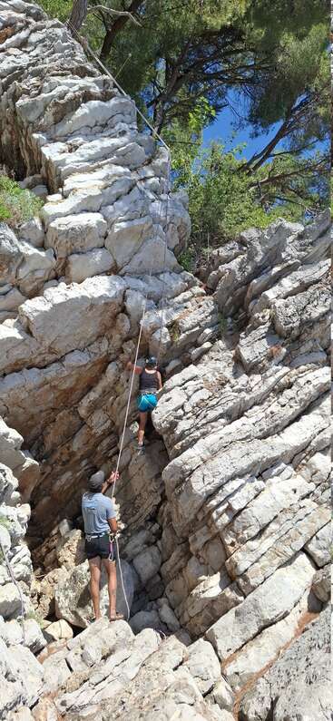 Un enfant escalade une falaise rocheuse abrupte tandis qu'un adulte l'assure depuis le bas. Tous deux portent un harnais et sont entourés de rochers et d'arbres escarpés sous un ciel bleu.