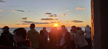Un grupo de personas se reúne junto al océano para contemplar una hermosa puesta de sol. El cielo está salpicado de nubes, lo que crea un ambiente tranquilo y pintoresco.