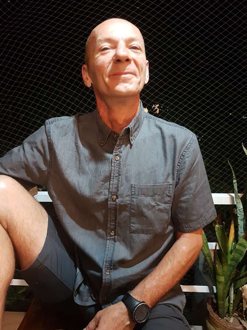 The image depicts a man sitting on a bench, wearing a gray shirt and shorts, with a watch on his left wrist, set against a dark background with a chain-link fence and plants.