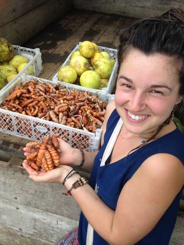 The image depicts a woman holding turmeric roots in her left hand, with crates of turmeric and other produce behind her, set against a wooden background.