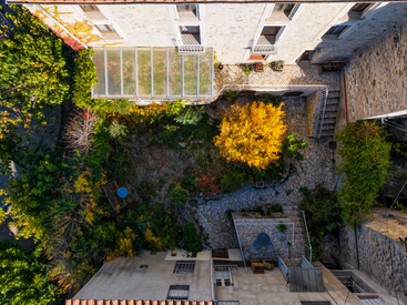 Diese Luftaufnahme zeigt einen bezaubernden Innenhof aus Stein, umgeben von Grünflächen und bunten Herbstbäumen, mit Gartenmöbeln, Wegen und Treppen, die zu den verschiedenen Wohnungseingängen führen.
