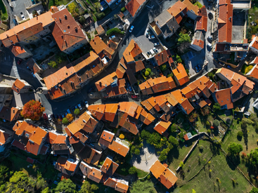 Eine Luftaufnahme eines Dorfes mit dicht gedrängten orangefarbenen Ziegeldächern, verwinkelten Gassen, geparkten Autos, viel Grün und umliegenden Feldern, die ein malerisches und lebendiges Bild des Dorflebens ergeben.