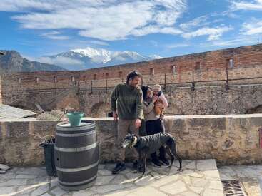 Eine Familie steht gemeinsam an einer Steinmauer, neben ihnen ein Hund. Hinter ihnen sind Berge, blauer Himmel und ein altes Backsteingebäude zu sehen. Ein friedlicher Moment.