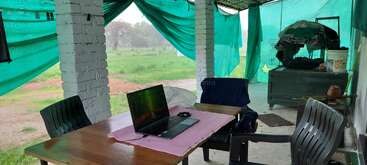 Ein Laptop steht auf einem Holztisch mit Plastikstühlen in einer offenen, grünbedeckten Veranda. Die Umgebung ist rustikal, mit einer üppigen, regnerischen Landschaft draußen.