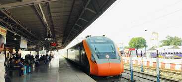 Ein moderner orangefarbener Zug steht an einem belebten Bahnsteig. Die Menschen warten und sitzen mit ihrem Gepäck. Die Bahnsteiguhr zeigt 14:51 Uhr an. Das Wetter scheint klar zu sein.