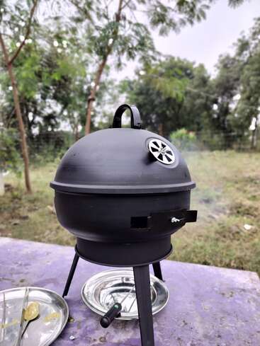 Ein kleiner schwarzer Holzkohlegrill steht auf einem lilafarbenen Tisch im Freien, umgeben von Bäumen und Grünzeug. In der Nähe sind Metallplatten und Zangen zu sehen.