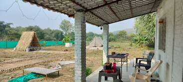 Dieses Bild zeigt eine ländliche Veranda mit Plastikstühlen und -tischen, mit Blick auf einen Hof mit einer strohgedeckten Hütte, einem Zelt, Pflanzen, Bäumen und einem grünen Zaun.