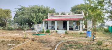 Ein kleines, weißes Backsteinhaus mit rotem Dach ist umgeben von Bäumen und offenem Land. Einfache Stühle, Gartenwege und Grünpflanzen schmücken den Hof.