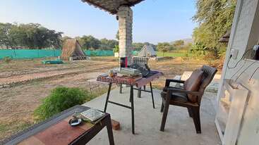A peaceful outdoor workspace features a desk with books, a typewriter, and chairs on a porch, overlooking a rural campsite with tents, greenery, and clear sky.