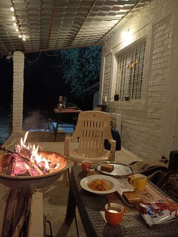 A cozy porch scene at night features a fire pit, snacks, tea, toasted sandwiches, a plastic chair, string lights, and a peaceful, relaxed atmosphere under a roof.