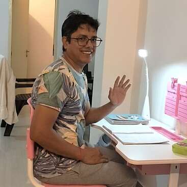 A smiling man with glasses, wearing a printed t-shirt and gray pants, sits at a desk with books, waving, illuminated by a desk lamp.