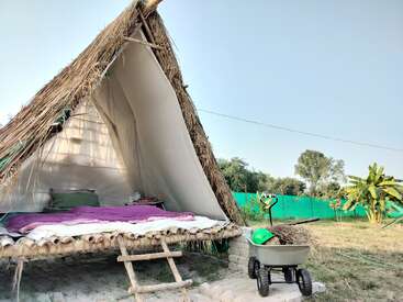 A rustic, triangular thatched hut with an open front reveals a cozy bed inside. Outside, a wheelbarrow and green plants add to the serene, rural setting.