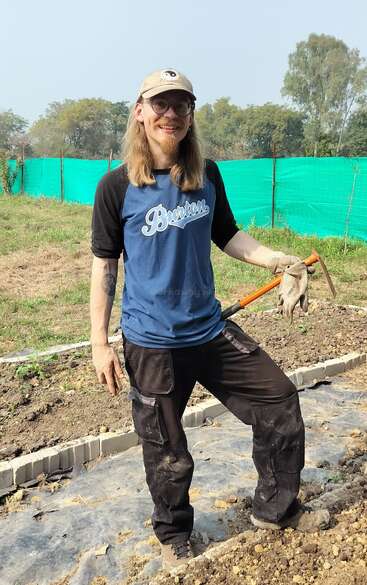 A man with long hair, glasses, and a cap stands in a garden. He is smiling, holding a gardening tool, dressed casually, and enjoying sunny outdoor work.