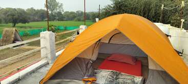 An orange tent is set up on a rooftop, containing a red mat, pillow, and blanket. Nearby fields and trees create a peaceful, rural outdoor scene.